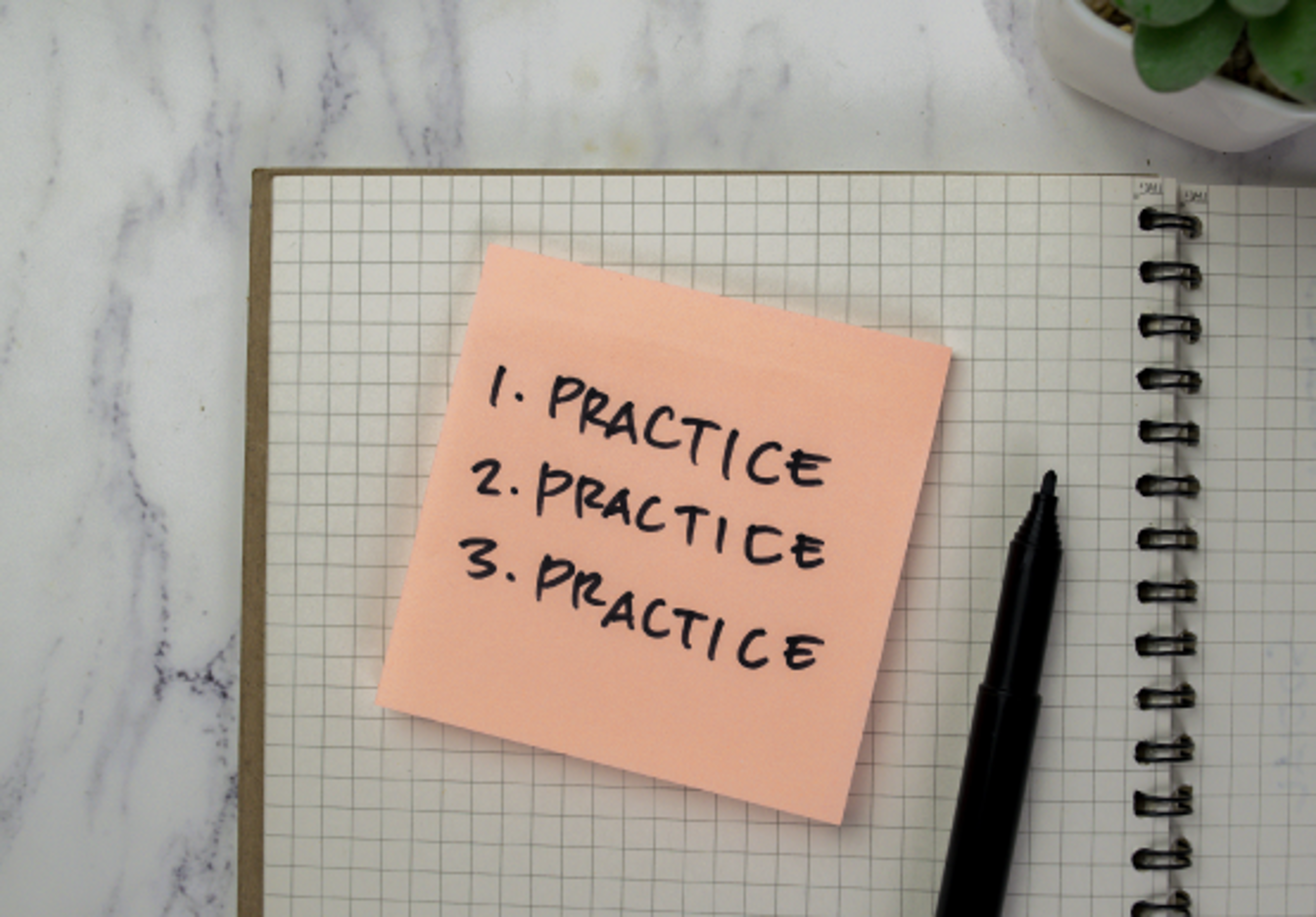 Grid lined notepad with small not saying 1. practice 2. practice 3. practice and a black pen to the right 